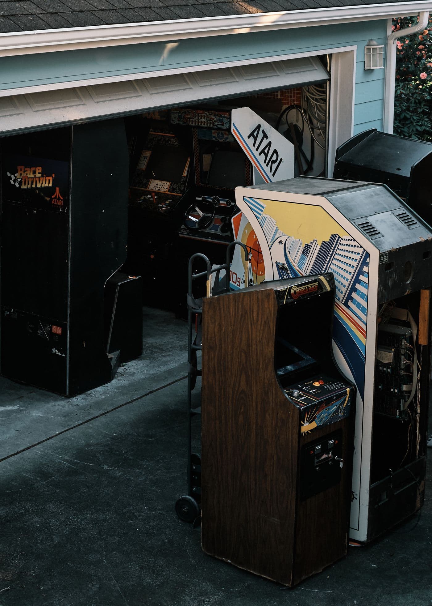 Vintage arcade cabinets gathered outside a garage, including wood-grain and racing machines
